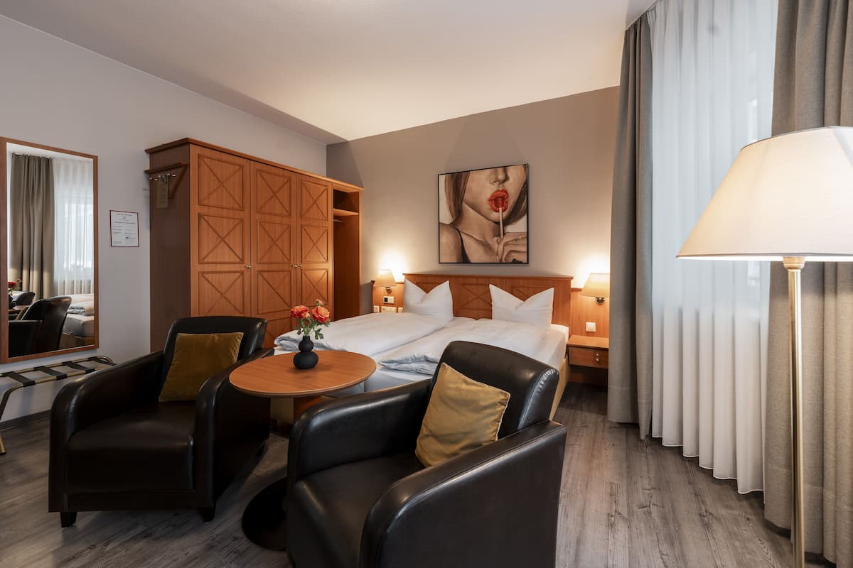 Ein Hotelzimmer mit Bett, zwei Stühlen, einem kleinen Tisch mit Blumen, einem Kleiderschrank und einem Bild an der Wand.