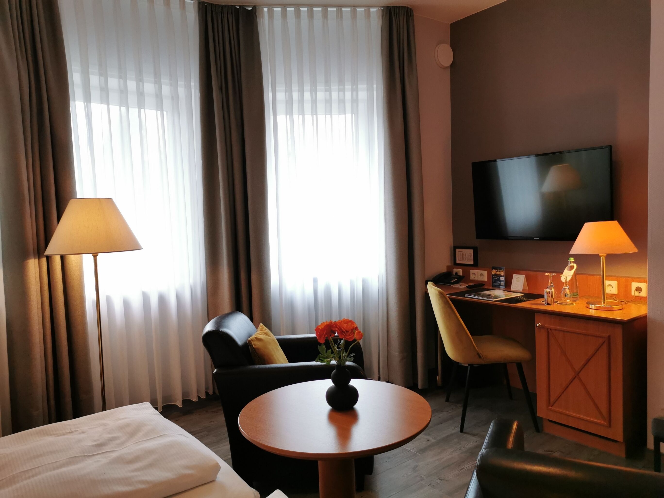 A hotel room with a bed, a round table with a vase of flowers, a desk with a chair, a television, and a lamp.