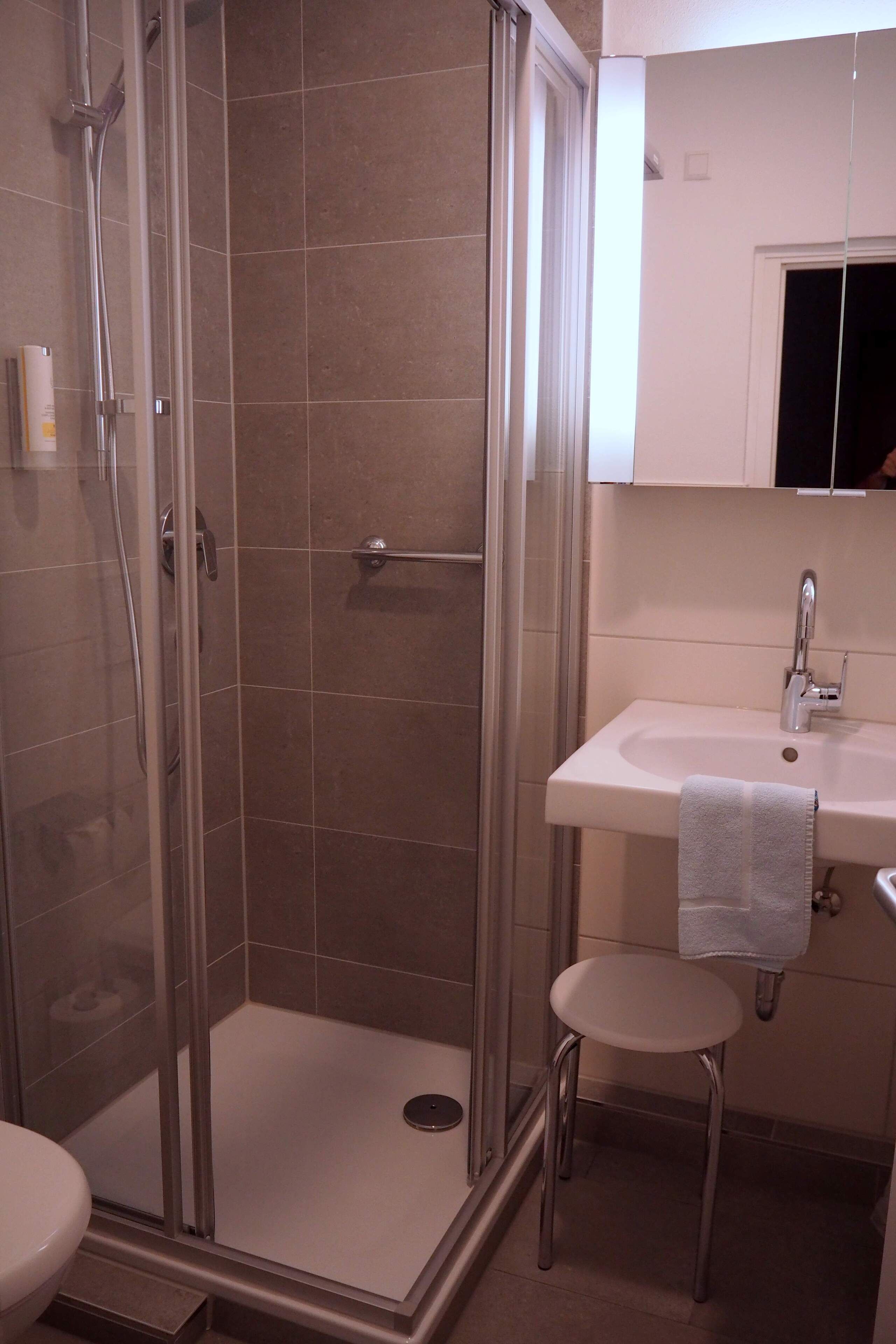 A bathroom with a glass-enclosed shower, a sink, and a small stool.