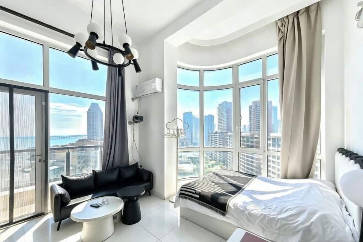 Une chambre d’hôtel moderne avec une grande fenêtre, un canapé noir, un lit blanc et une petite table ronde.