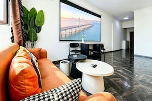 Un moderno vestíbulo de hotel con una pantalla grande mostrando un puente, un sofá con cojines naranjas, una mesa de centro y una planta en maceta.
