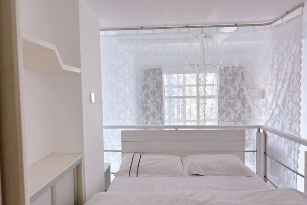 A bedroom with a bed, a nightstand, a window with lace curtains, and a white ceiling.