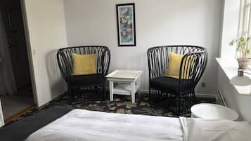 Una habitación con dos sillones de mimbre negro con cojines amarillos, una mesita blanca y una cama con ropa de cama blanca.