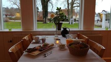 A breakfast table set with food, drinks, and a candle, overlooking a garden and buildings.