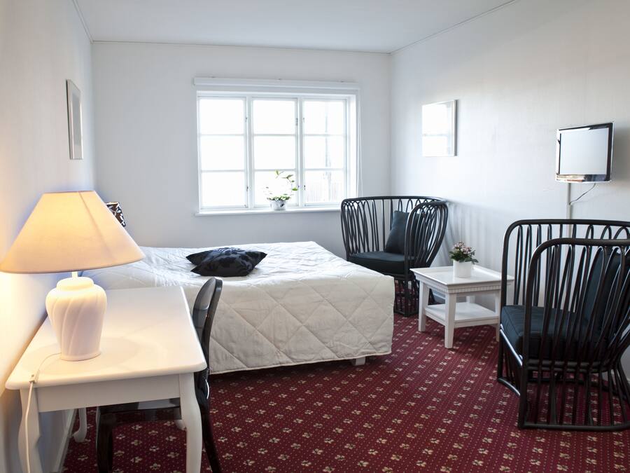 A hotel room with a bed, a desk with a lamp, a chair, a small table, and a TV.
