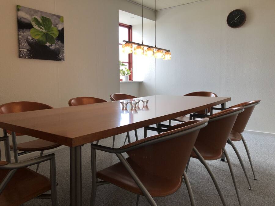 A conference room with a long table, six brown chairs, a clock on the wall, and a hanging light fixture.