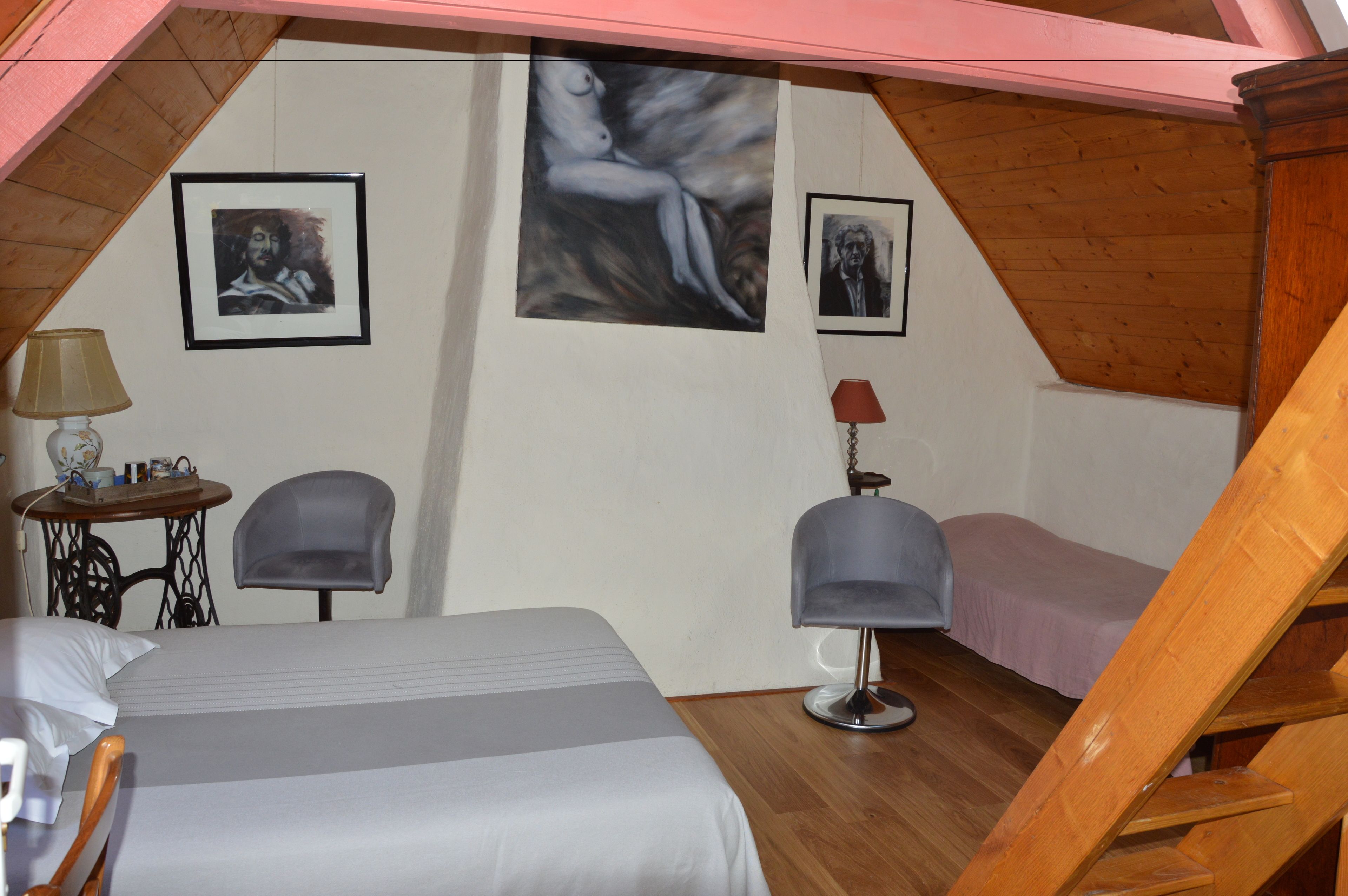 A bedroom with two beds, a chair, a table with a lamp, and artwork on the walls.