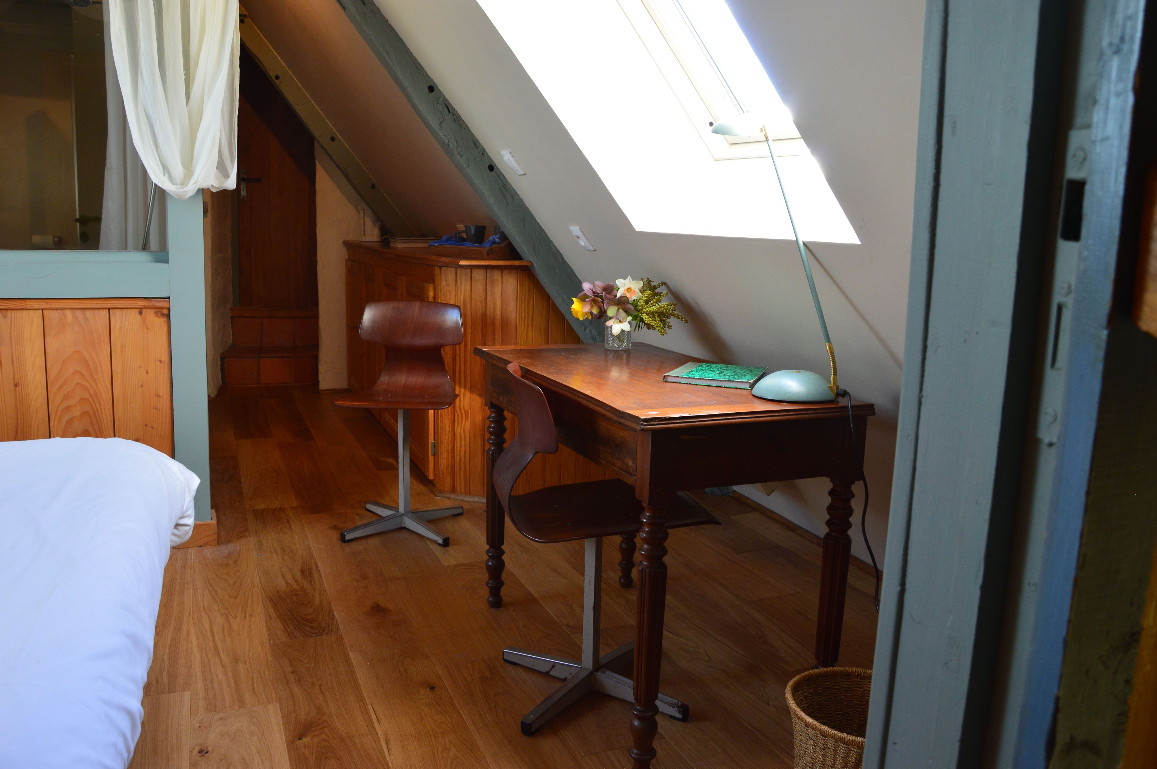 A bedroom with a wooden floor, a bed, a desk with a chair, a lamp, and a window with a curtain.