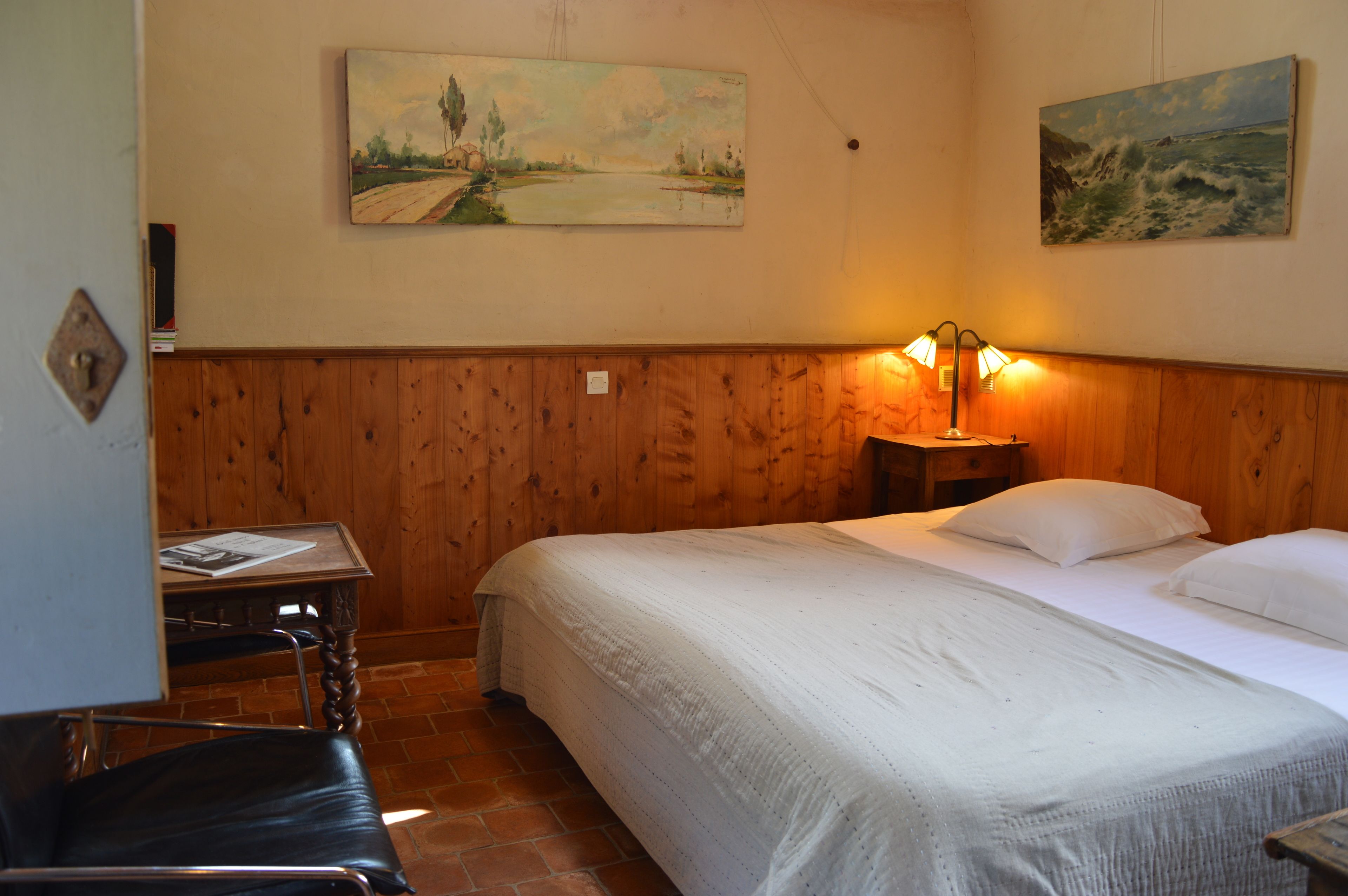 Una habitación con una cama, cabecera de madera, una mesita de noche con lámpara, una silla y dos cuadros en la pared.