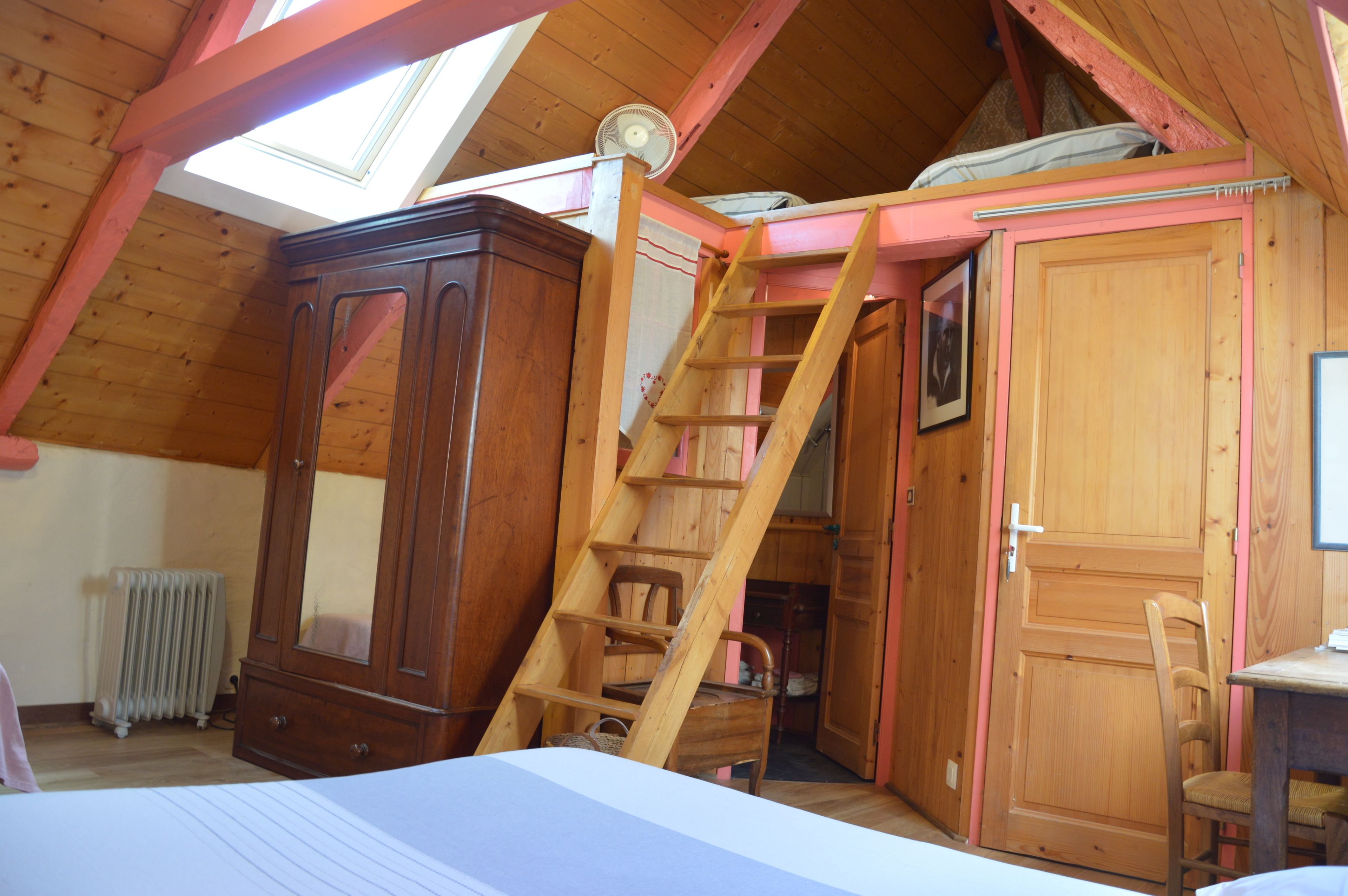 Een slaapkamer met een schuine kap, een houten garderobe, een ladder, een radiator en een bureau met stoel.
