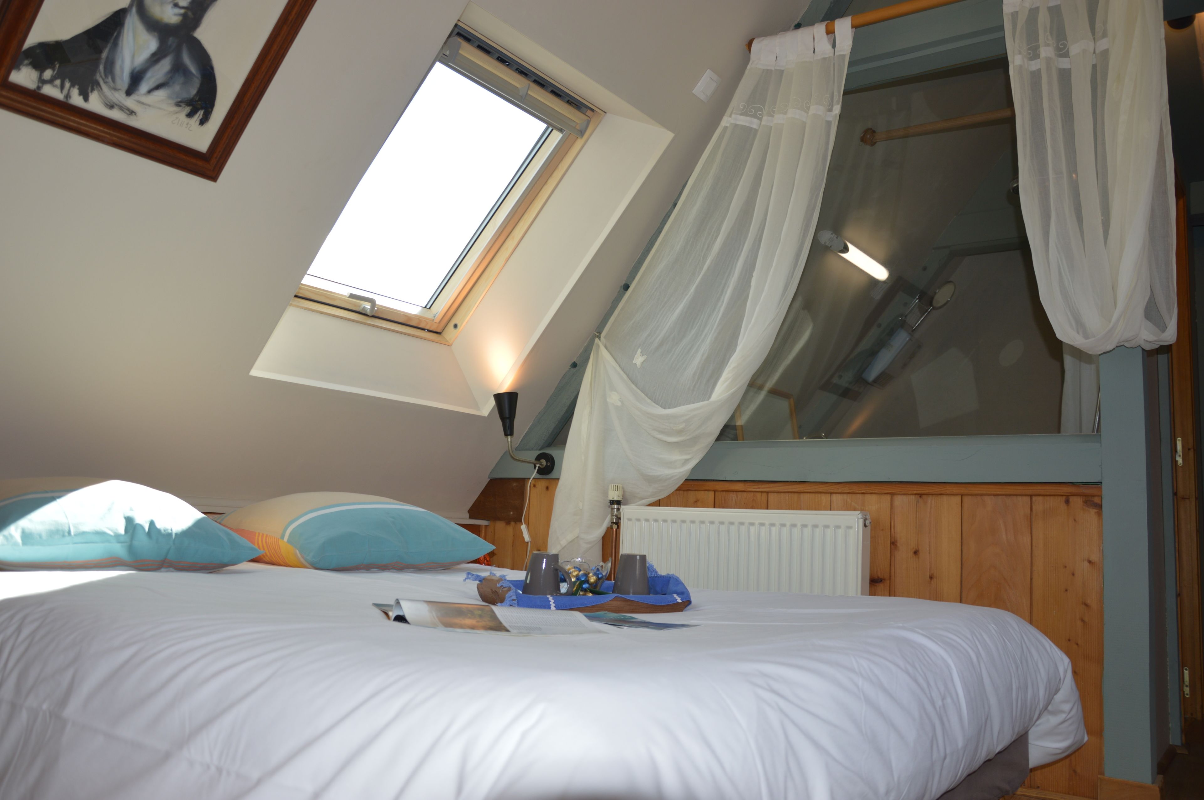 Een slaapkamer met een schuine kap, een groot raam met dunne gordijnen, een bed met witte beddengoed en een houten hoofdbord.