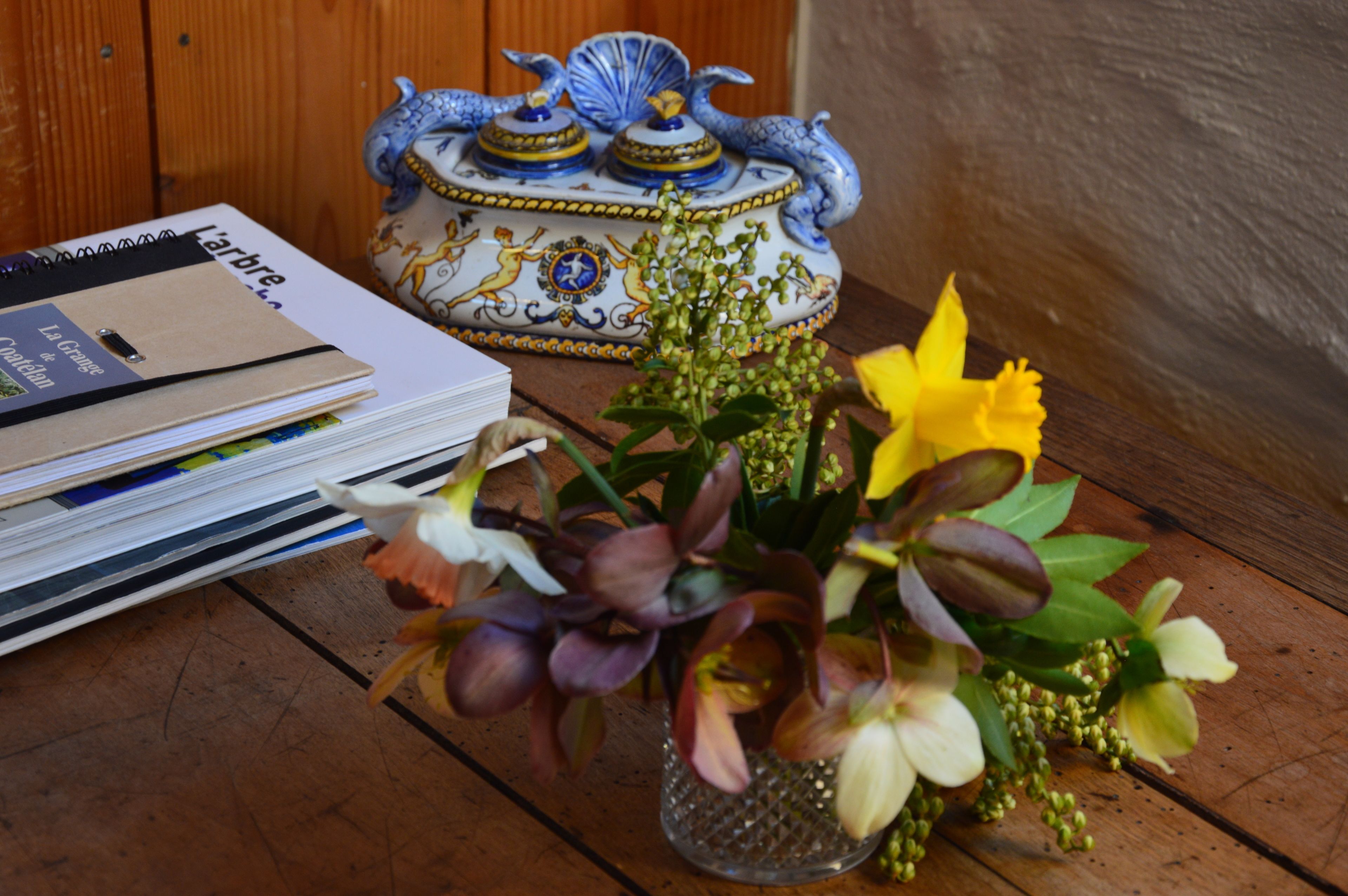 Une table en bois sur laquelle se trouvent un pot en céramique décoratif, une pile de cahiers et un petit vase avec des fleurs.