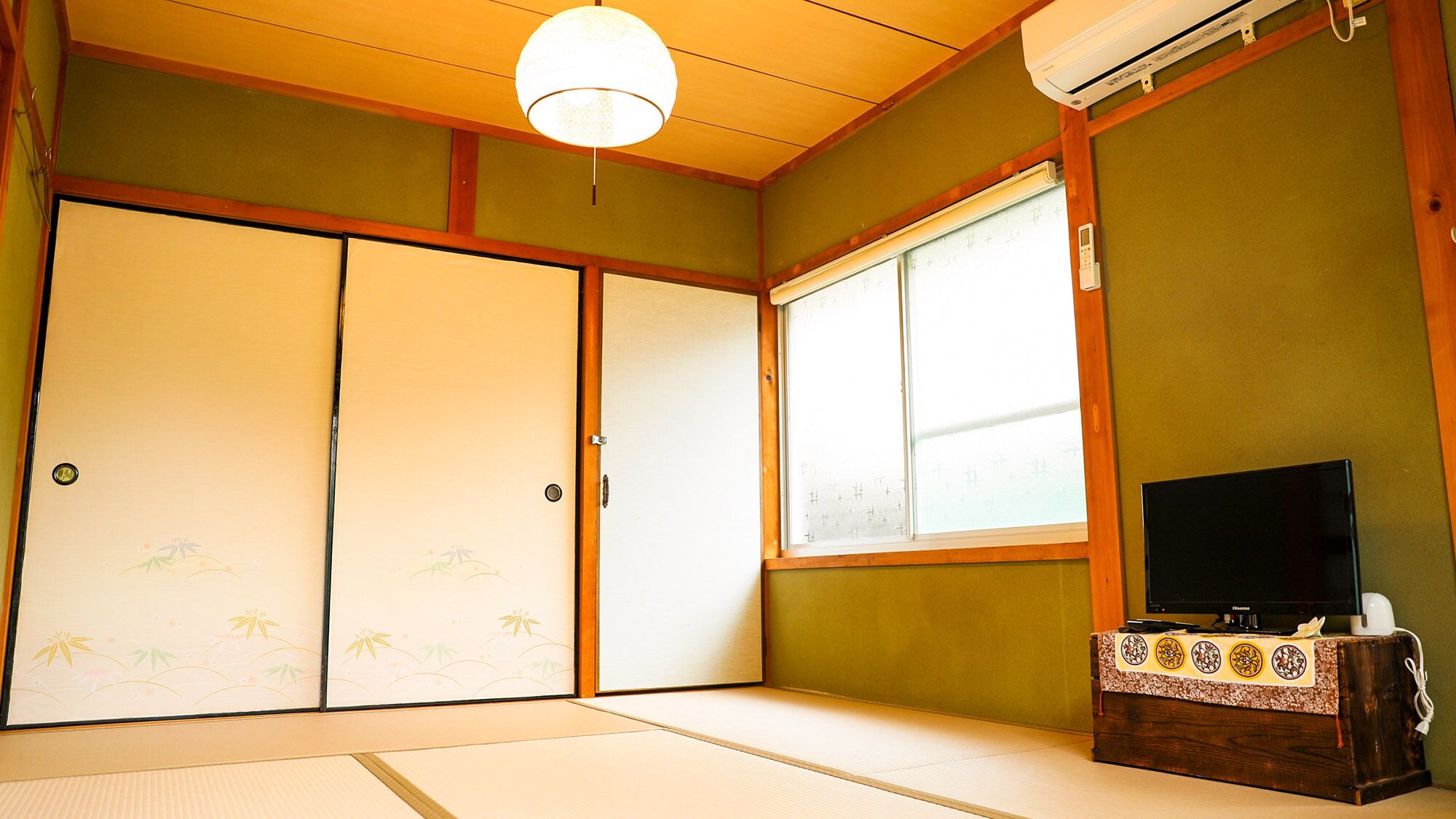 A traditional Japanese room with tatami flooring, sliding doors, a television on a wooden stand, and an air conditioning unit.