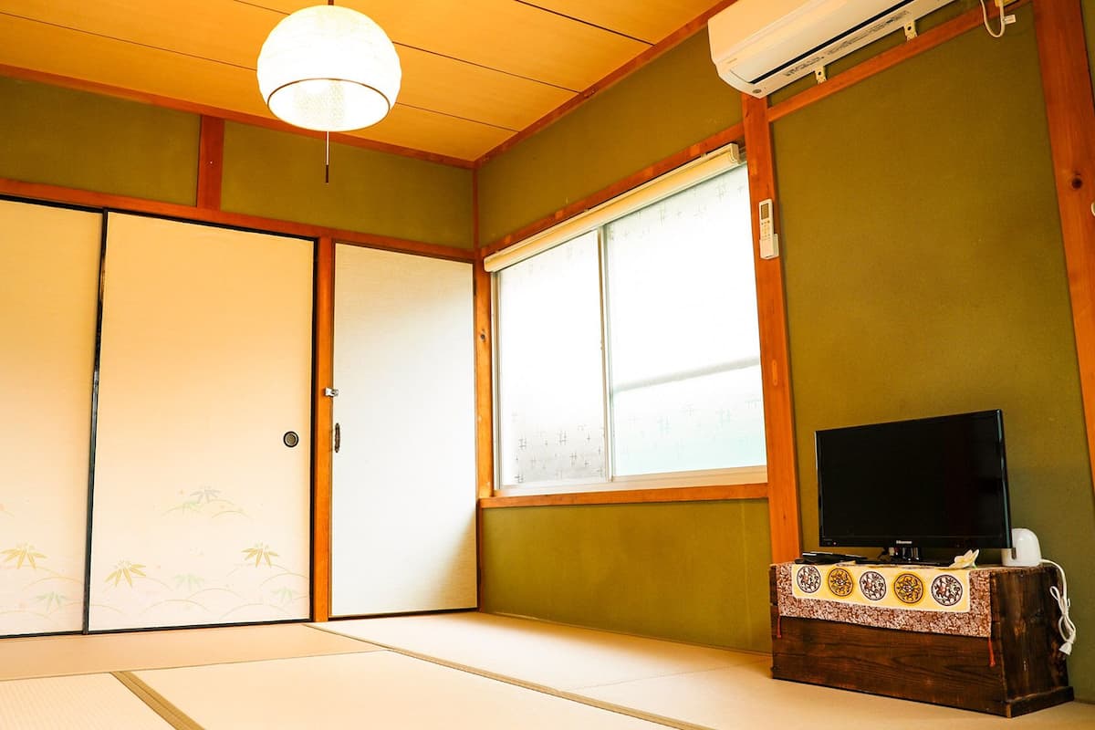 Ein traditioneller japanischer Raum mit Tatami-Boden, Schiebetüren, einem Fernseher auf einem Holzbodenständer und einer Klimaanlage.