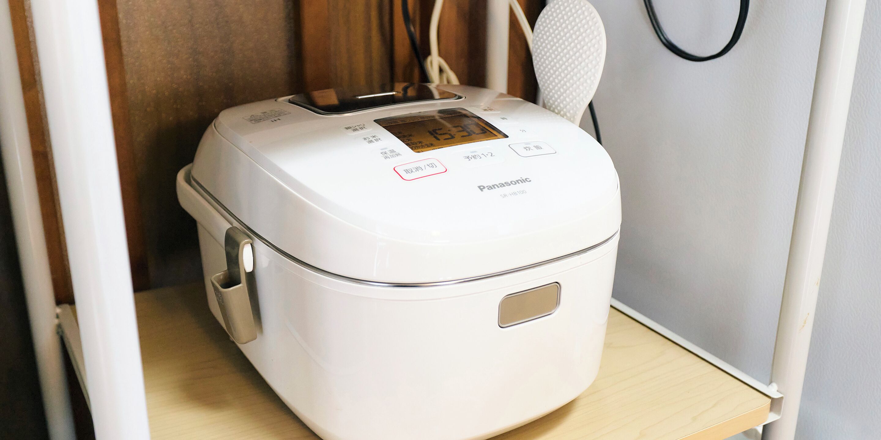 A white Panasonic rice cooker with a digital display, placed on a wooden shelf.