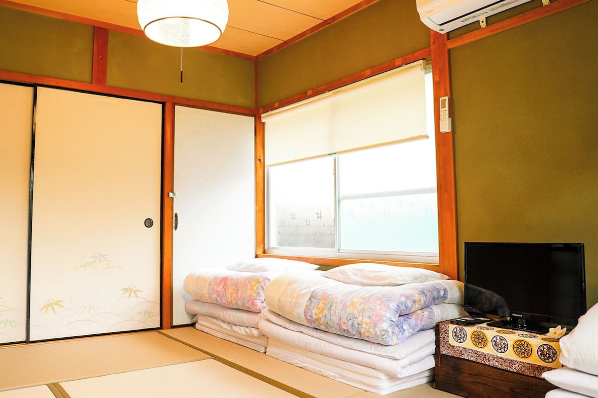 Ein traditioneller japanischer Raum mit Tatami-Boden, Futon-Bett, Schiebetüren und einem großen Fenster.