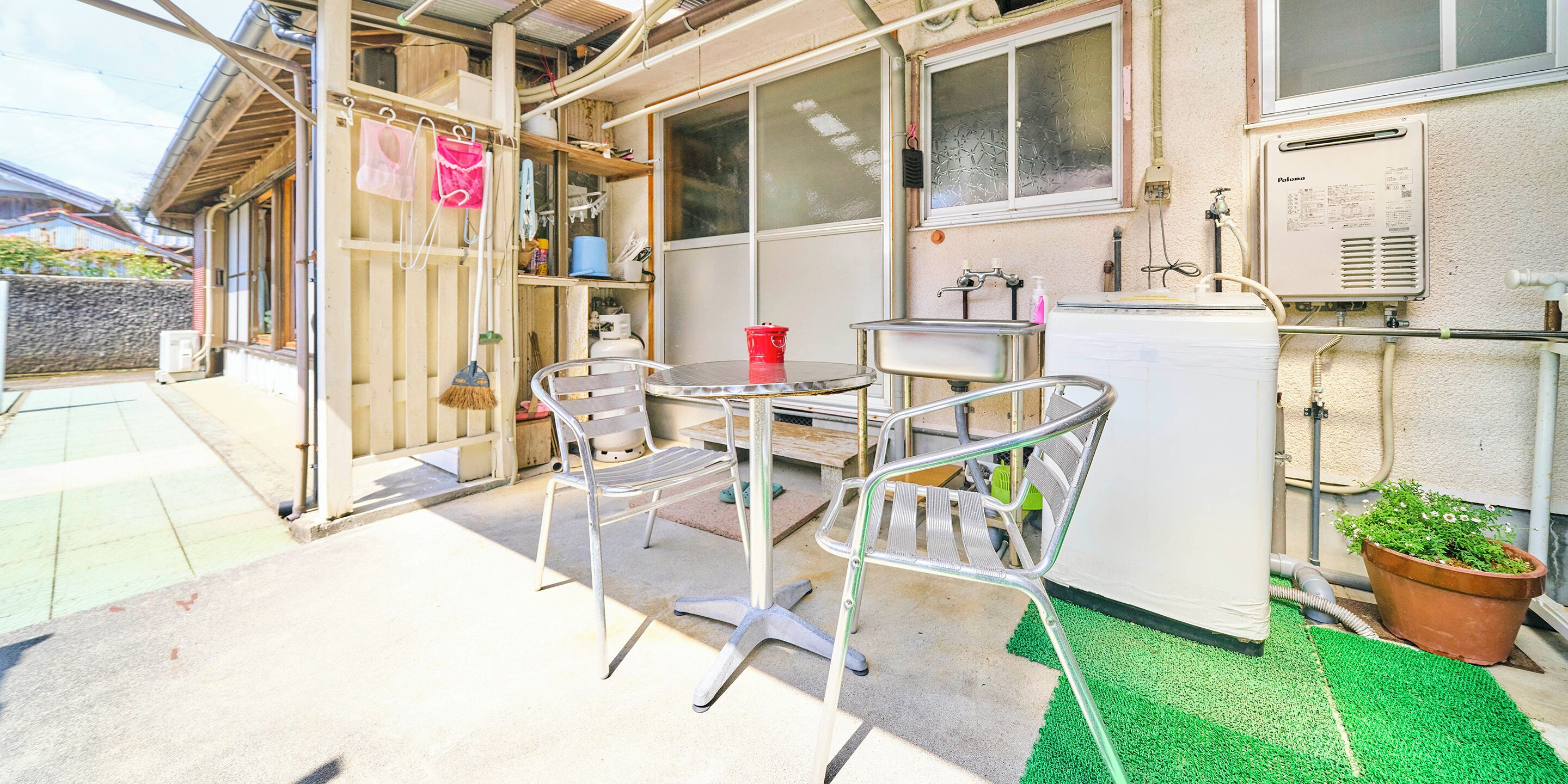 A small outdoor area with a metal table and chairs, a red bucket, a white refrigerator, and a large potted plant.