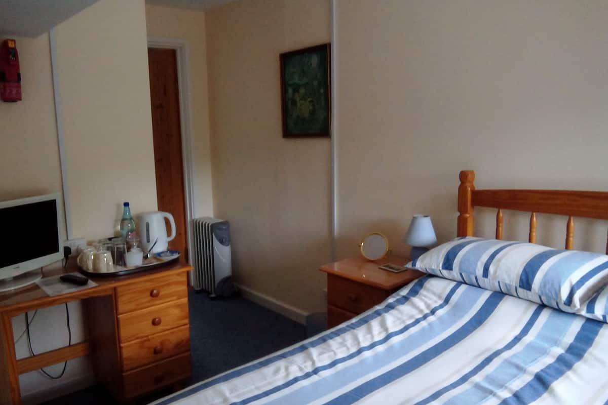 A hotel room with a bed, a desk with a computer, a radiator, a mirror, and a bedside table with a lamp.