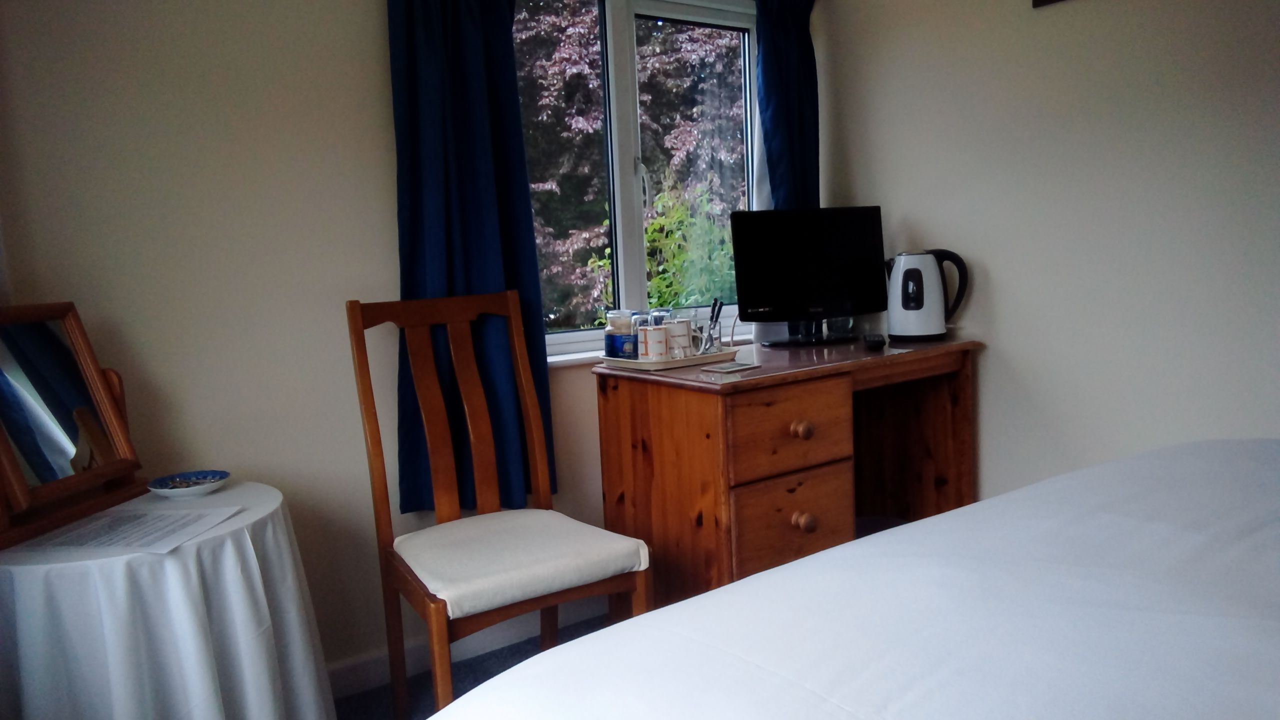 A bedroom with a bed, a wooden desk with a computer and a kettle, a chair, a small table with a bowl, and a window with curtains.