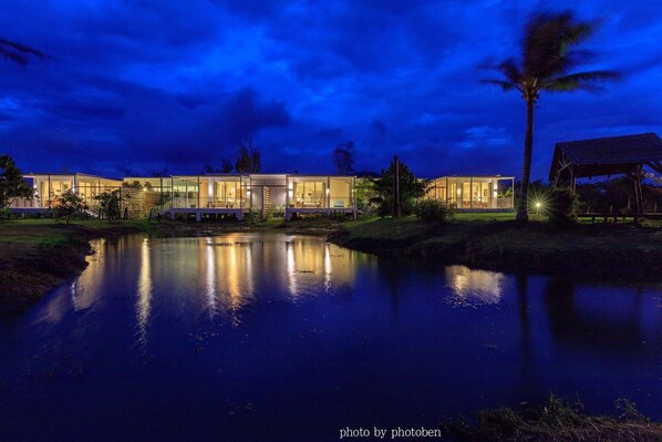 Un hotel moderno con una piscina reflectante al frente, rodeado de exuberante vegetación y palmeras.