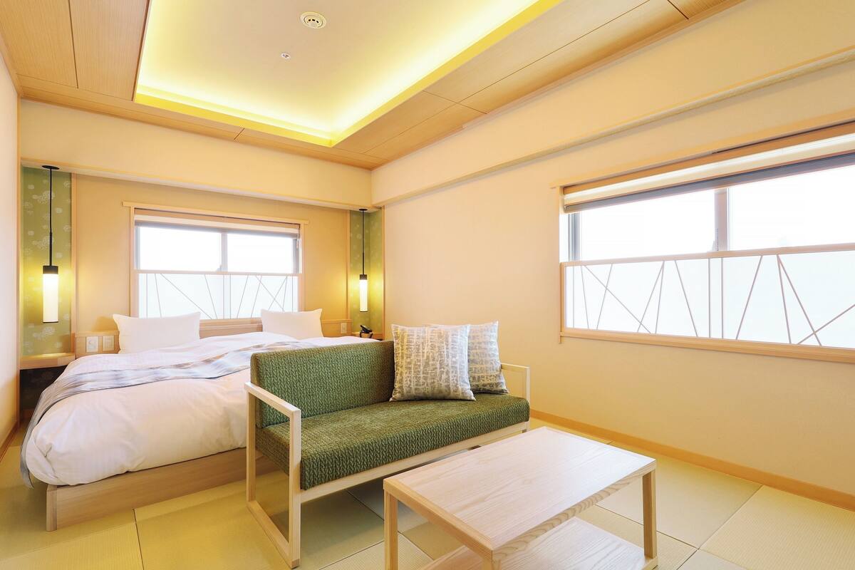 Quarto de hotel moderno com uma cama, um sofá verde, uma mesa pequena e janelas amplas.
