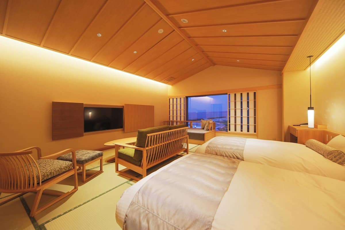 Quarto de hotel com uma cama grande, televisão, área de refeições com mesa e cadeiras, e vista para o mar através de uma janela ampla.