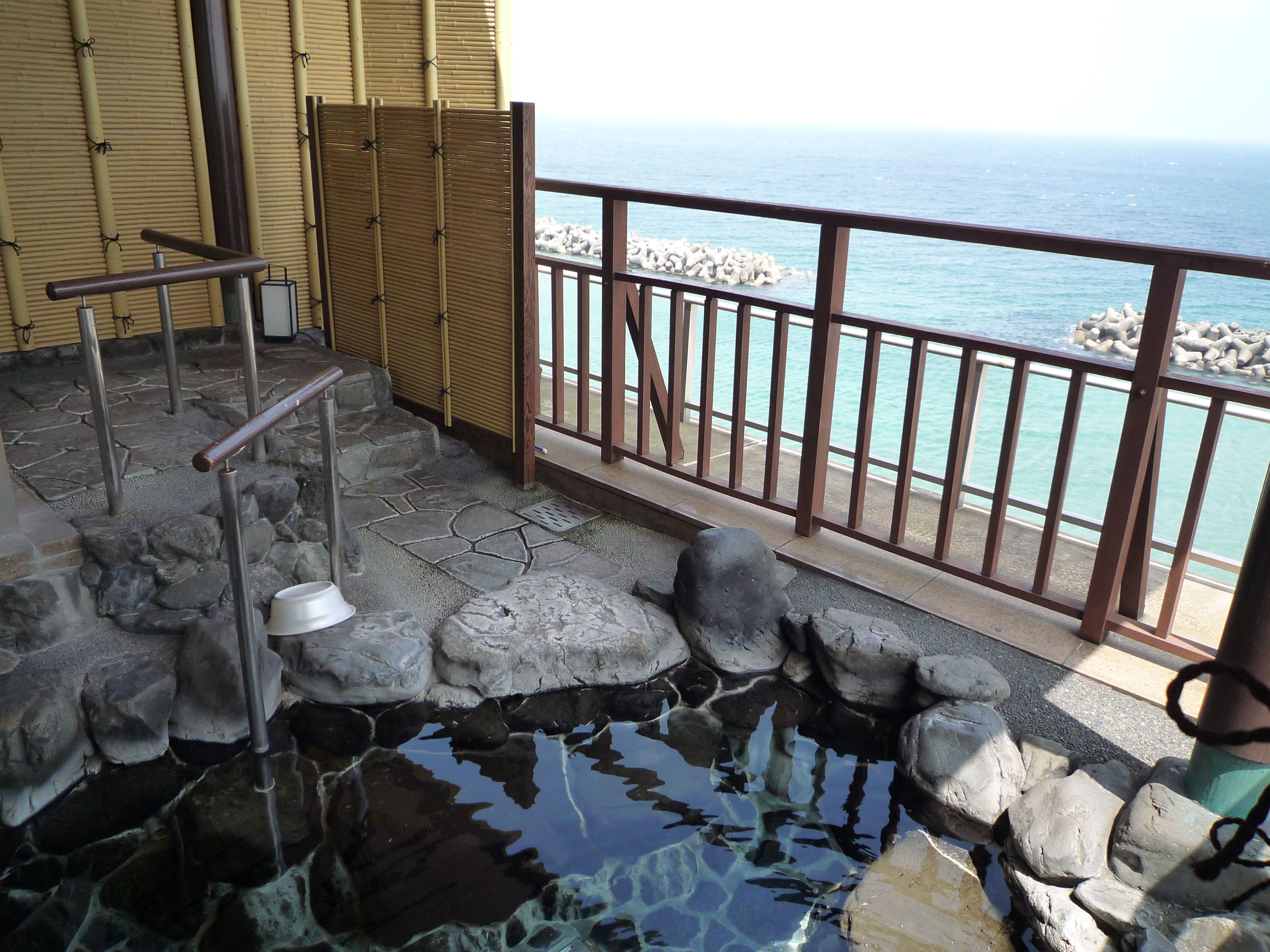 Una piscina de piedra con cubierta de madera, cerca de una valla de bambú, y vista al mar.