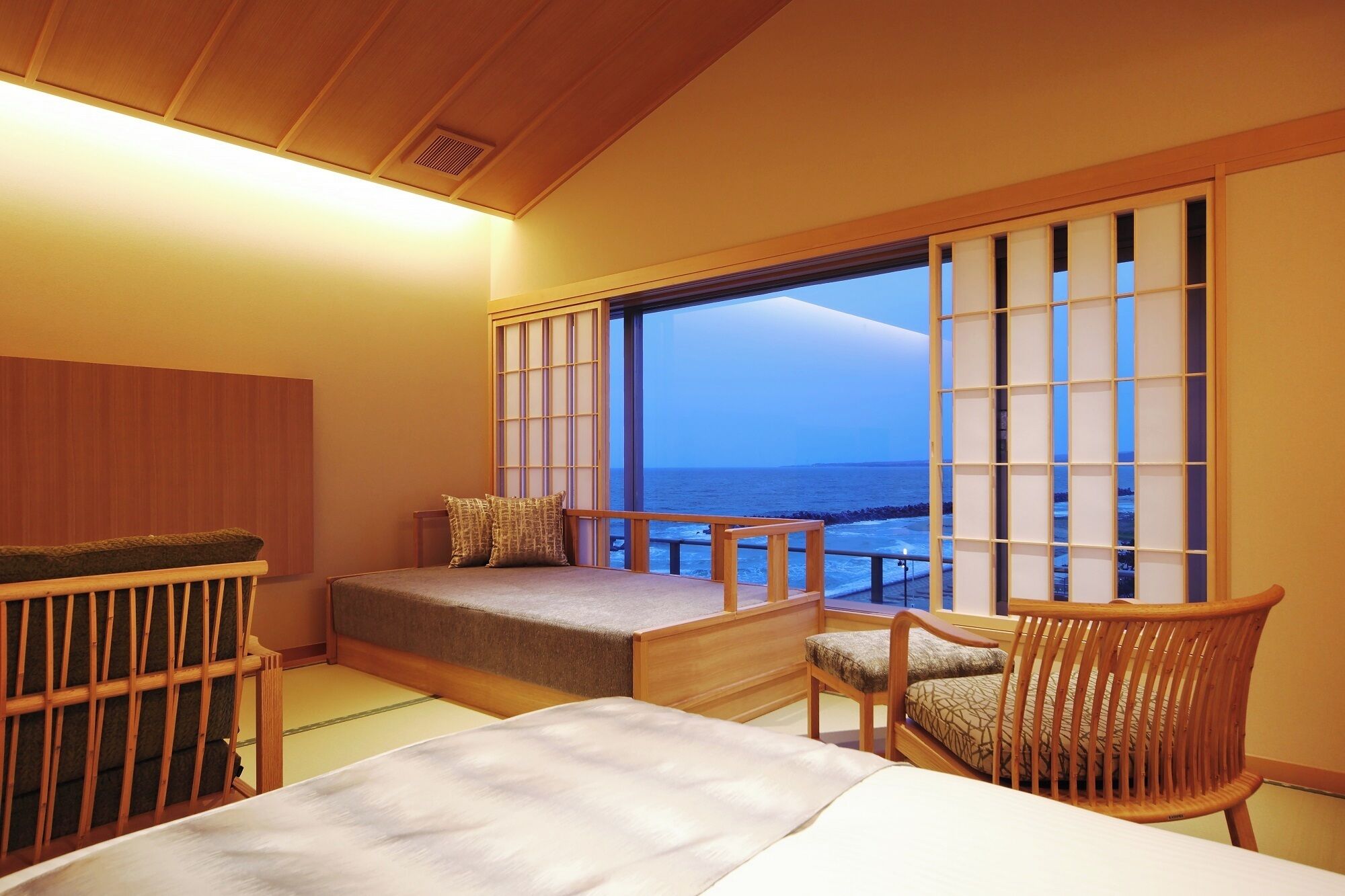 A room with a bed, a chair, and a sofa. There is a large window with a view of the sea.