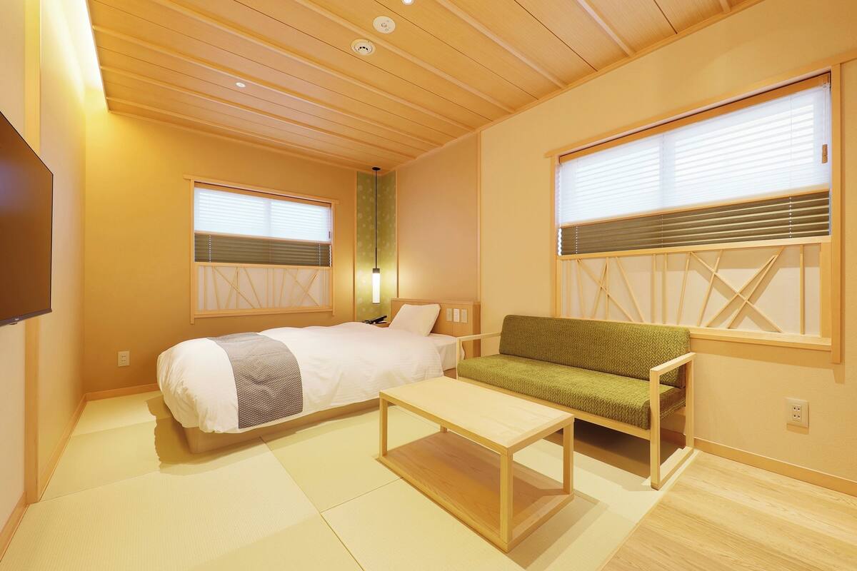 Um quarto com uma cama, um sofá verde, uma mesa de madeira e duas janelas com persianas.