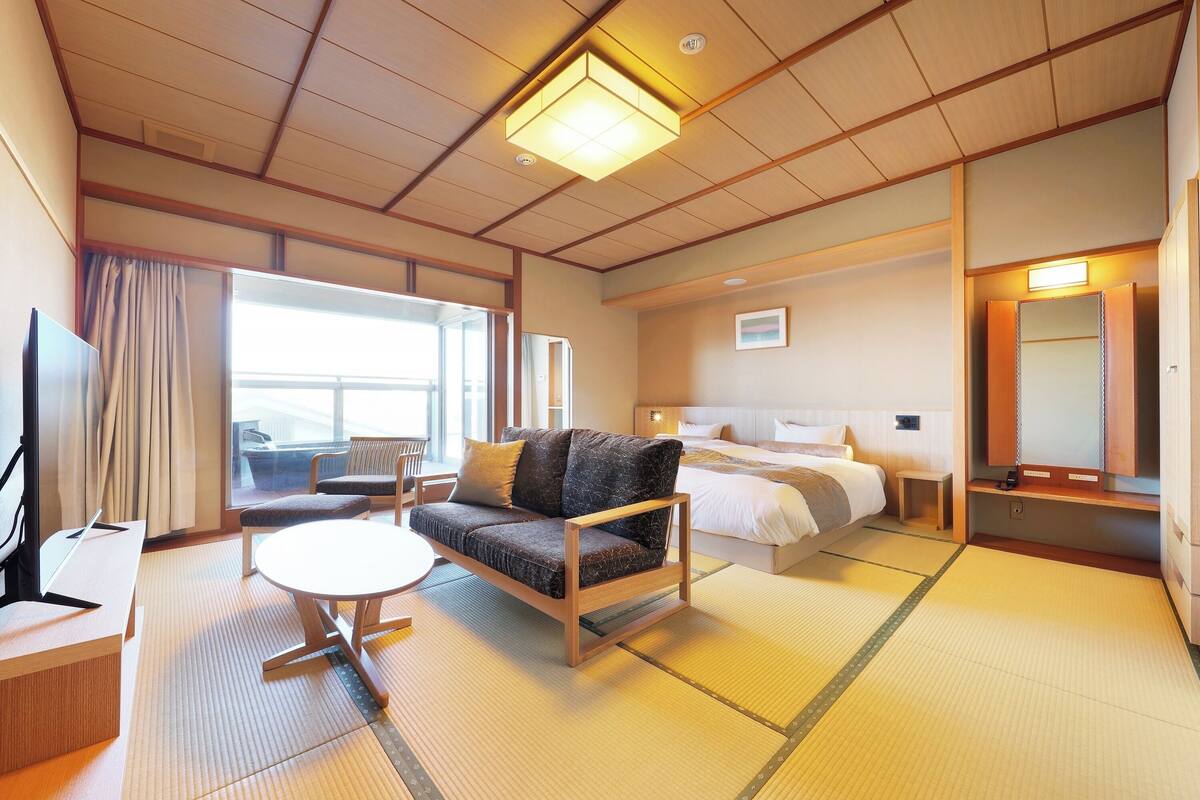 Quarto de hotel com sofá, mesa redonda pequena, cama e uma janela grande.