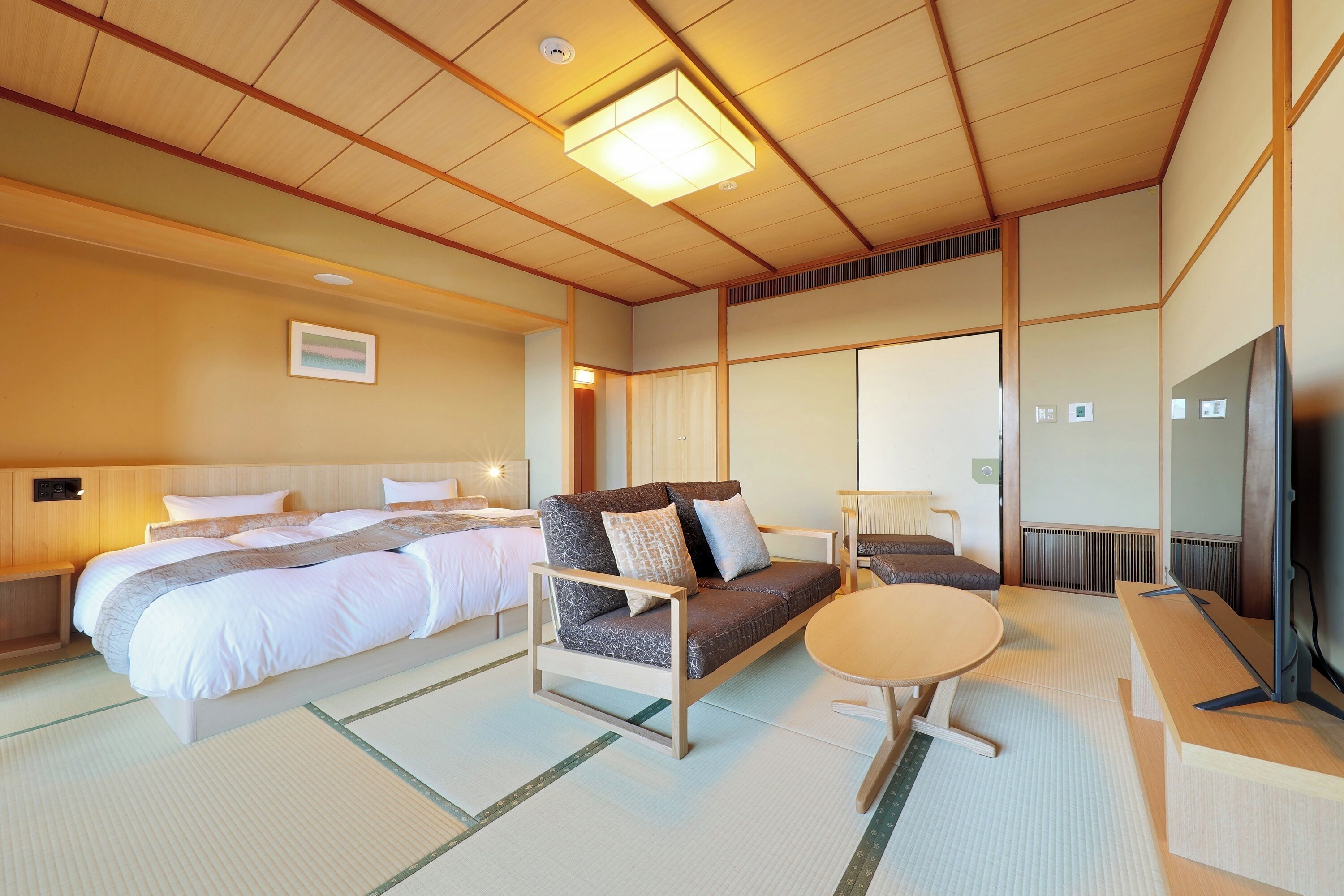A traditional Japanese-style room with tatami flooring, a bed with white bedding, a sofa with cushions, a small round table, and a television.