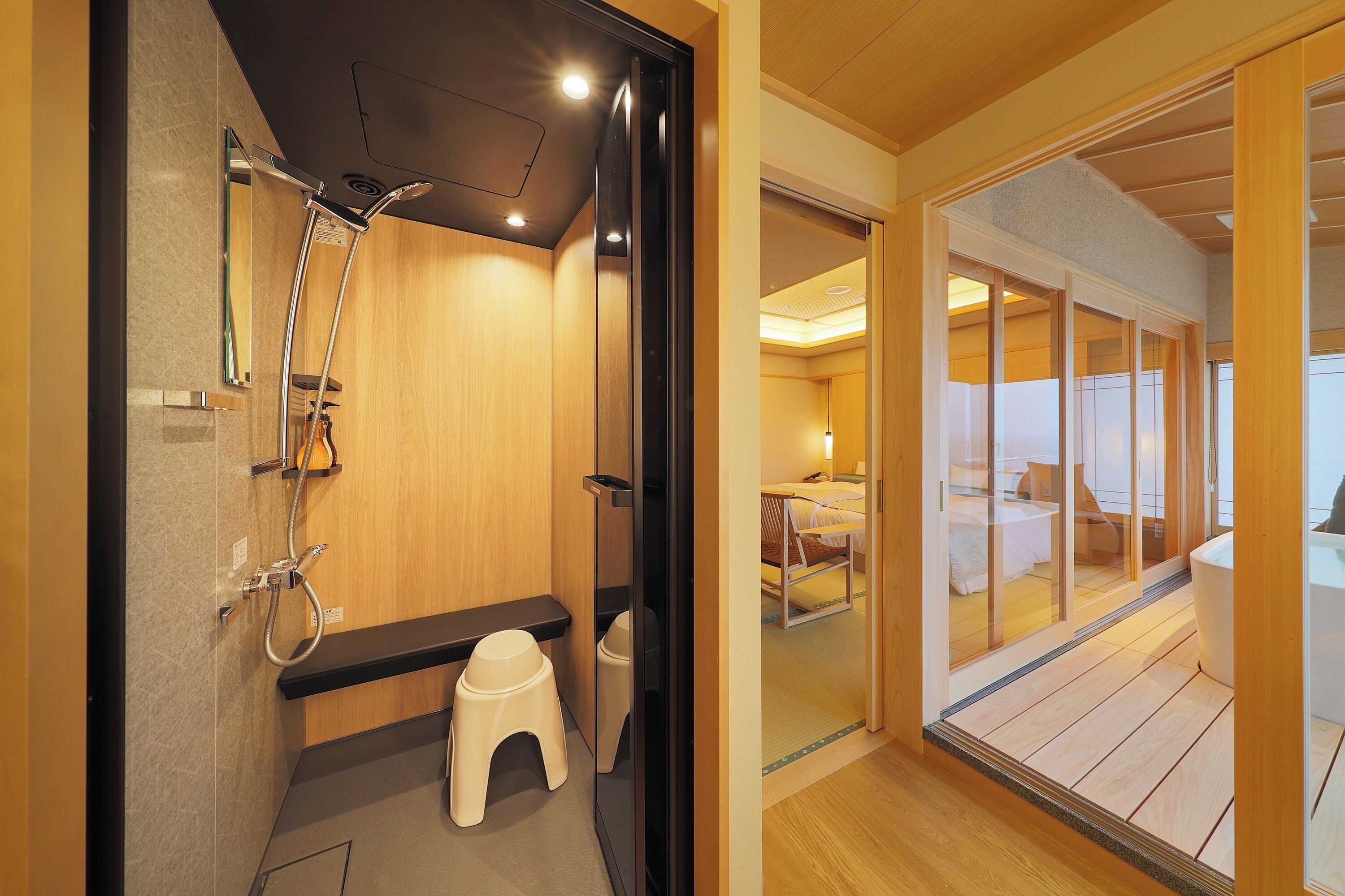 A modern bathroom with a shower, toilet, and a small stool. It features wooden walls and a visible bedroom with a bed and window.