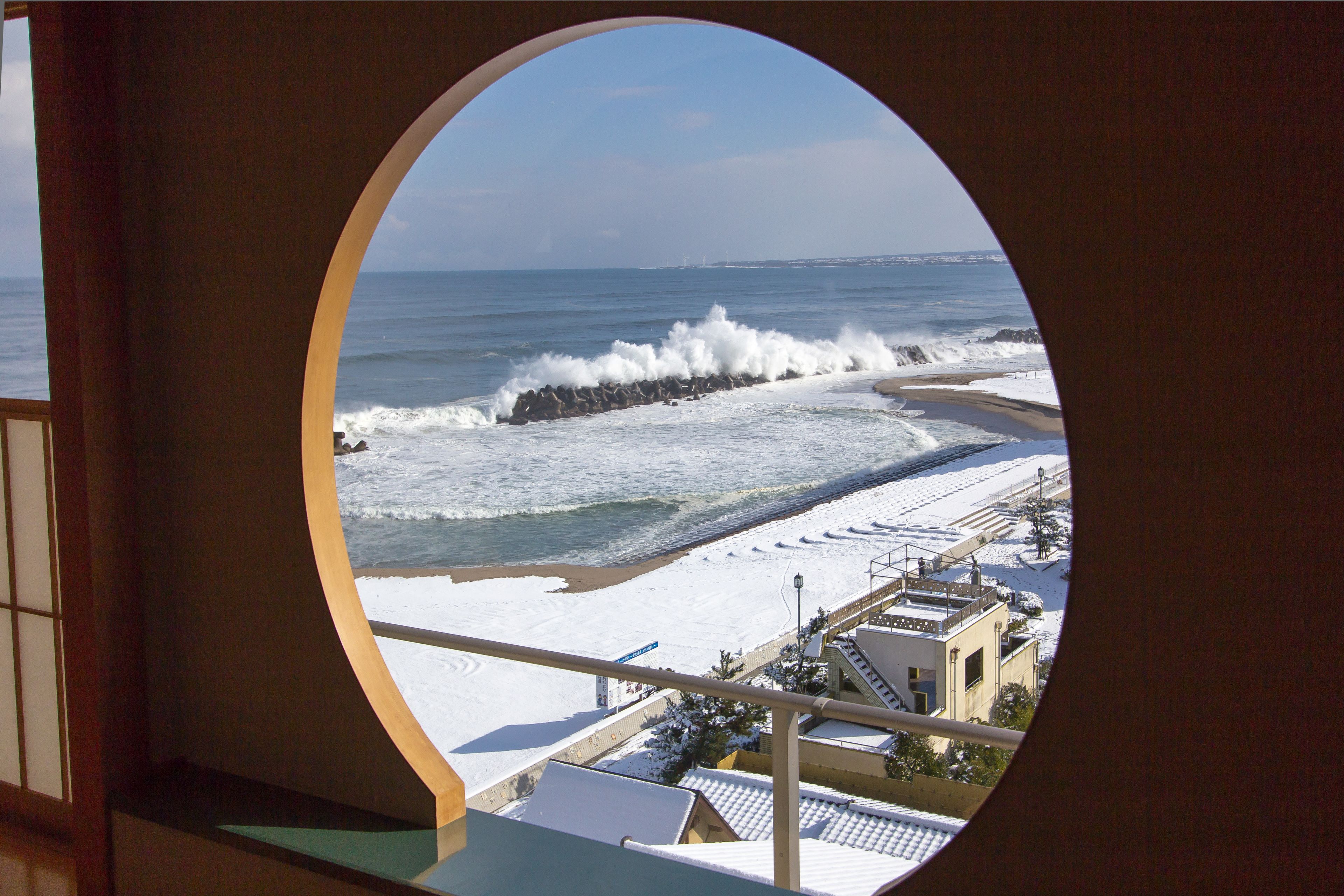 A view through a circular window of a beach with waves crashing onto the shore, a sandy beach, and buildings in the background.
