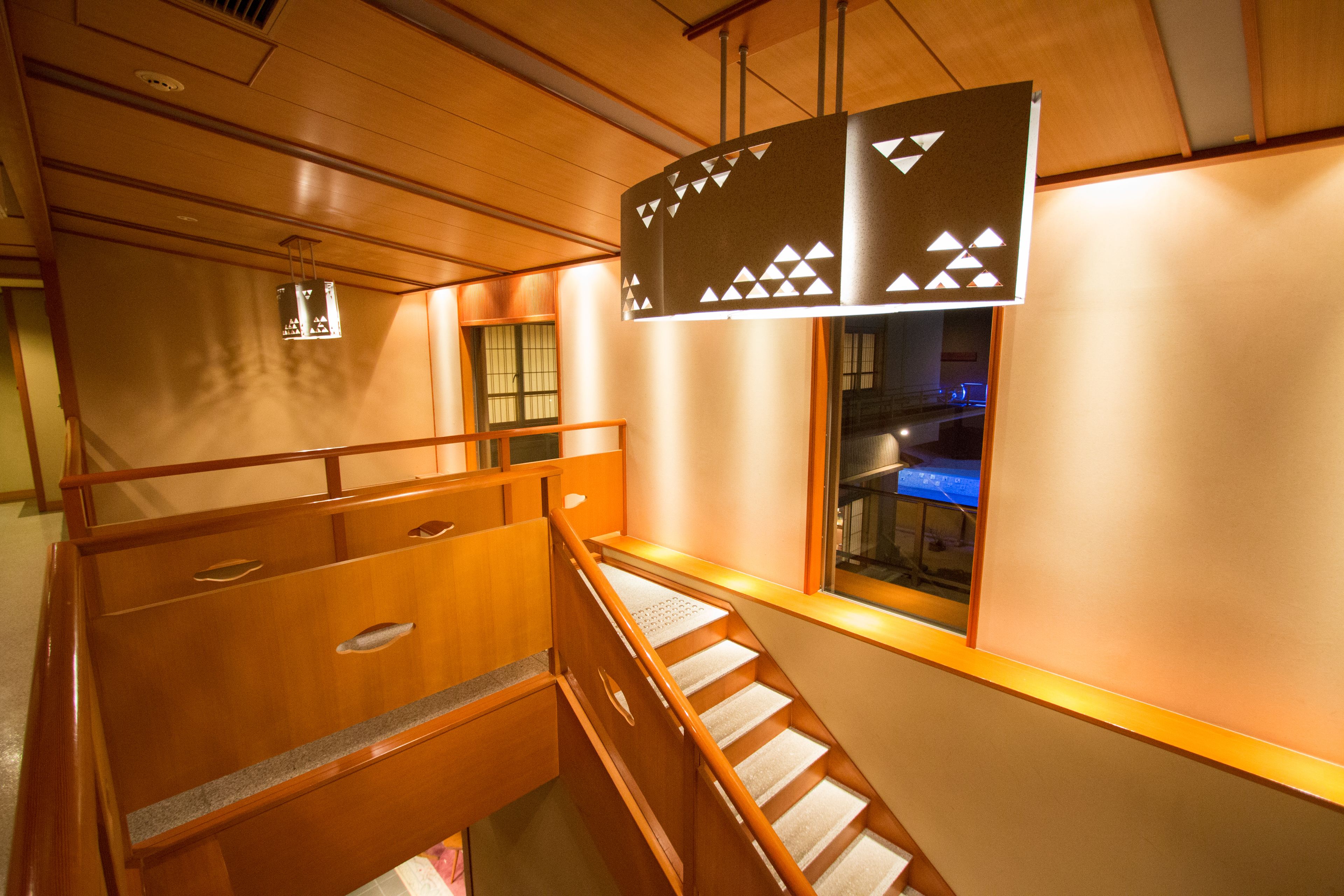 Un intérieur en bois avec un escalier, un luminaire suspendu et une fenêtre donnant sur une pièce éclairée d’une lumière bleue.