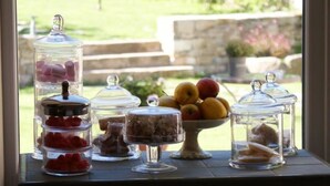 A table with various food items in glass containers, a bowl of fruit, and a window view of a garden. - Les Fréjals (Compeyre)