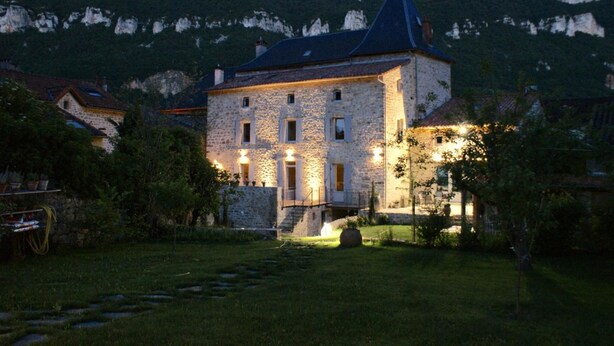 A stone building with a steep roof and a balcony, surrounded by a well-maintained garden and a mountainous backdrop. - Les Fréjals (Compeyre)