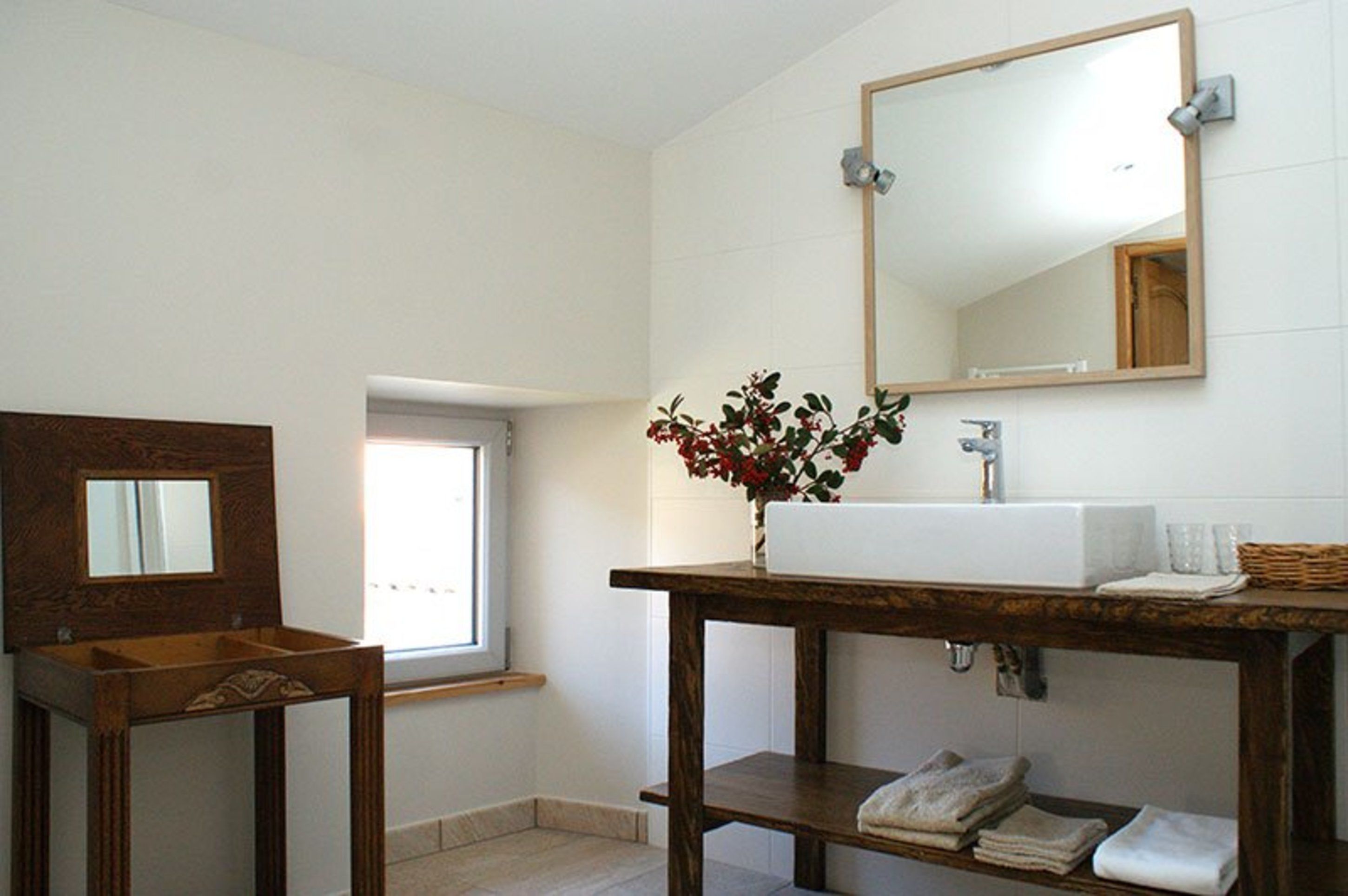 A bathroom with a wooden vanity, a white sink, a mirror, and a vase with red flowers.