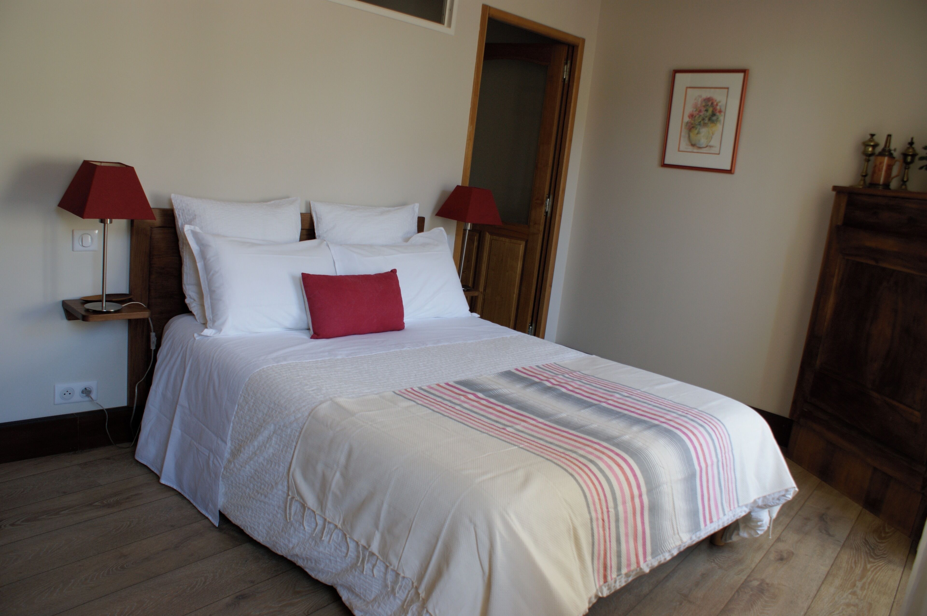 Una cama bien hecha con ropa de cama blanca y un almohadón rojo, con dos lámparas rojas a cada lado sobre mesitas de noche de madera.