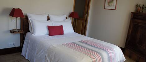 Una cama bien hecha con ropa de cama blanca y un almohadón rojo, con dos lámparas rojas a cada lado sobre mesitas de noche de madera.