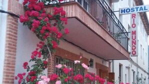 Une auberge dotée d’un balcon orné de fleurs rouges, avec un mur de pierre en premier plan et une enseigne indiquant le nom de l’auberge.