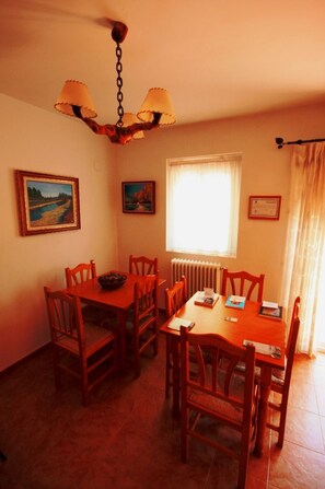 Een eetruimte met een houten tafel en stoelen, een kroonluchter, ingelijste foto’s aan de muur en een raam met gordijnen.