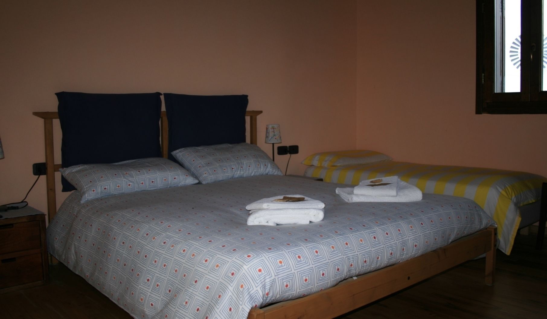A double bed with a patterned bedspread and two pillows, a bedside table with a lamp, and a window with blinds.