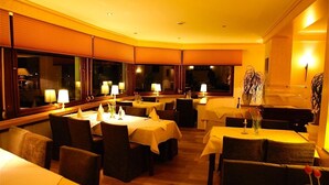 A restaurant with multiple tables and chairs, each with a white tablecloth. There are lamps on each table, and the room has large windows with blinds. - Moselhotel Restaurant Peifer (Brodenbach)