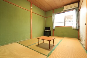 Et værelse med tatami-gulv, et lille træbord, et fjernsyn på et skab og et vindue med gardiner.