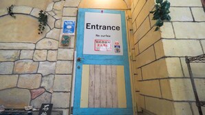 Ein Eingangsbereich mit einer Steinmauer, einer blauen Tür und einem Schild mit den Aufschriften 'Entrance' und 'No Curfew'.