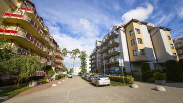 Ein Wohngebäude mit mehreren Balkonen, einer gepflasterten Straße, geparkten Autos und blauem Himmel.