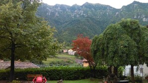 Une zone dallée avec des chaises rouges et un banc, entourée d’arbres, et offrant une vue sur un village et des montagnes en arrière-plan.