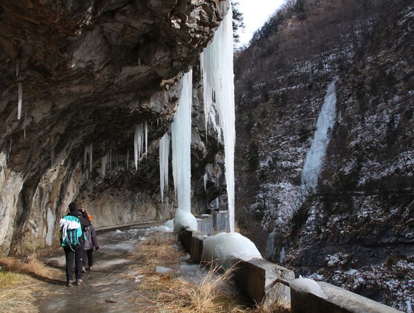 A narrow path flanked by rocky cliffs with icicles hanging from the ceiling and a waterfall on the right. - Ostello Angolo Verde (Angolo Terme)
