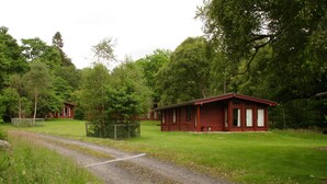 Un bâtiment de style rustique, semblable à un chalet, avec un chemin de gravier menant à l’entrée, entour é d’une végétation luxuriante et d’arbres.