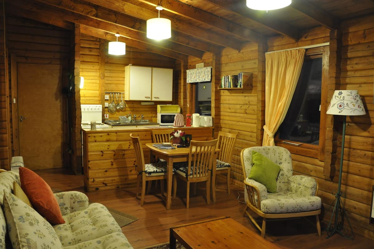 Ein gemütliches Blockhaus-Interieur mit einem Holzesstisch, Stühlen, einer Couch und einem Fenster mit Vorhängen.